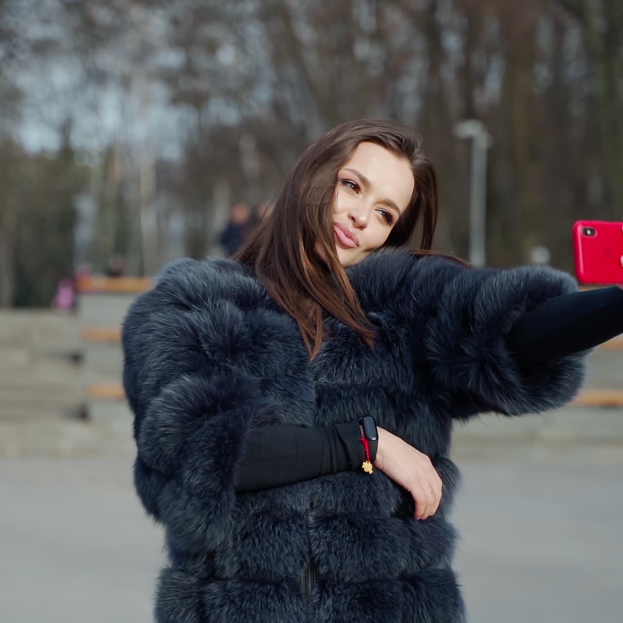 Portrait of a beautiful girl doing selfie in the park. Young woman in luxury fur coat doing photo of herself on the red phone in a sunny winter day.