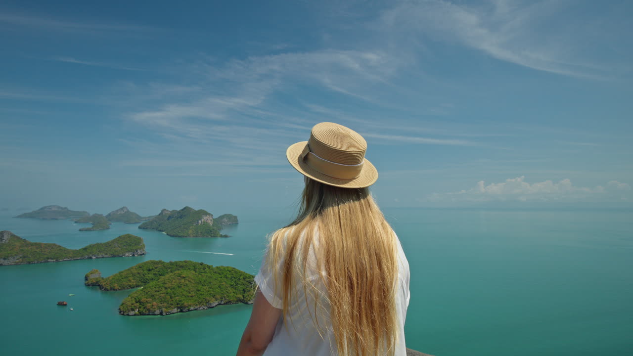 Jovem mulher com um chapéu de palha em um mirante alto, olhando para o mar turquesa e as exuberantes ilhas verdes do Parque Marinho Ang Thong na Tailândia, apreciando a paisagem de ilha tropical. Vídeos em 4K de alta qualidade.