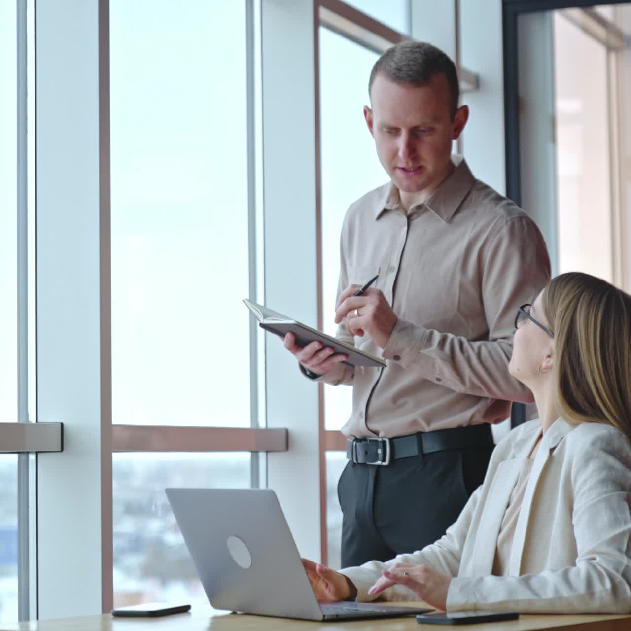 Colleagues have conversation at the table near the window. Woman employee sitting at computer and man standing over her