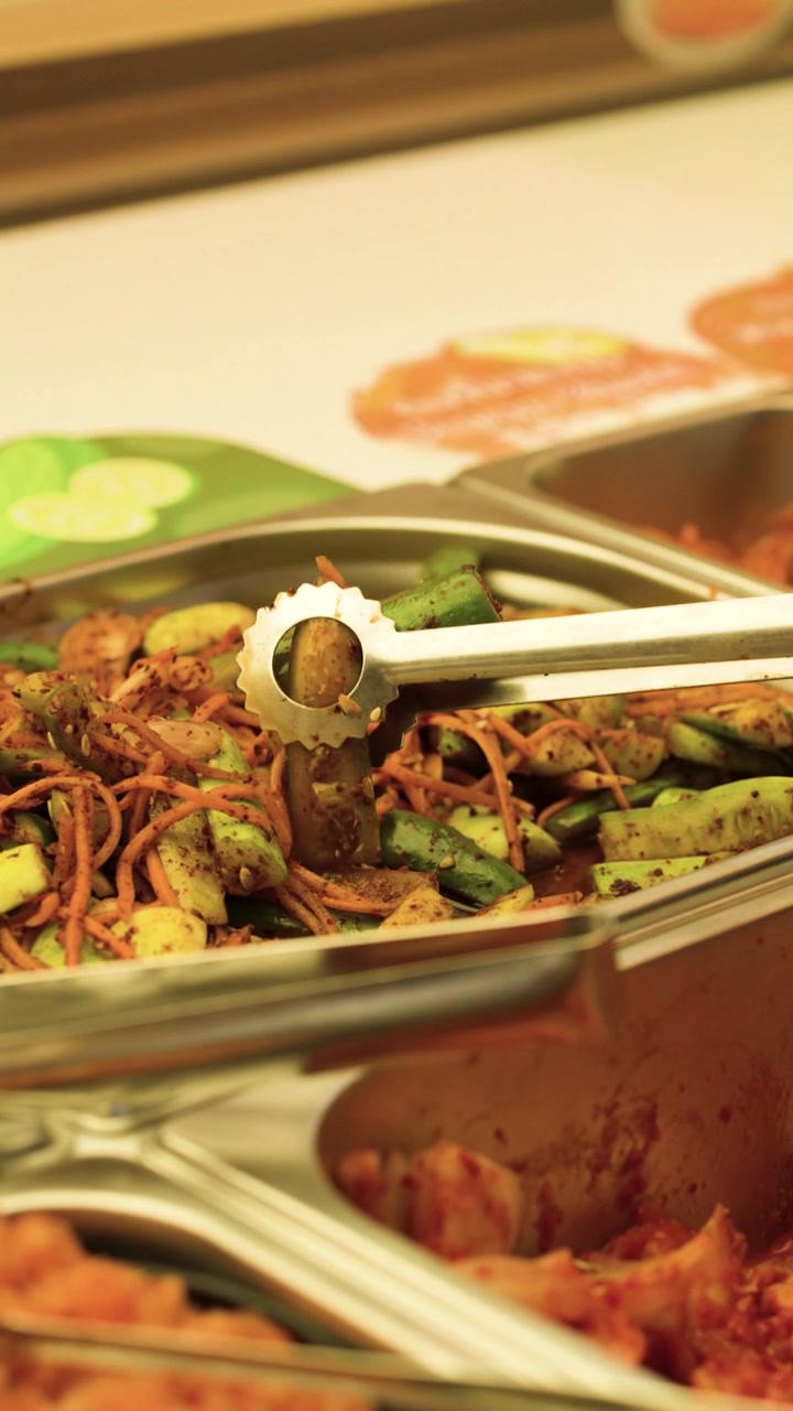 Pinze metalliche prendono kimchi piccante di cetrioli fermentati da un vassoio da buffet in acciaio inossidabile