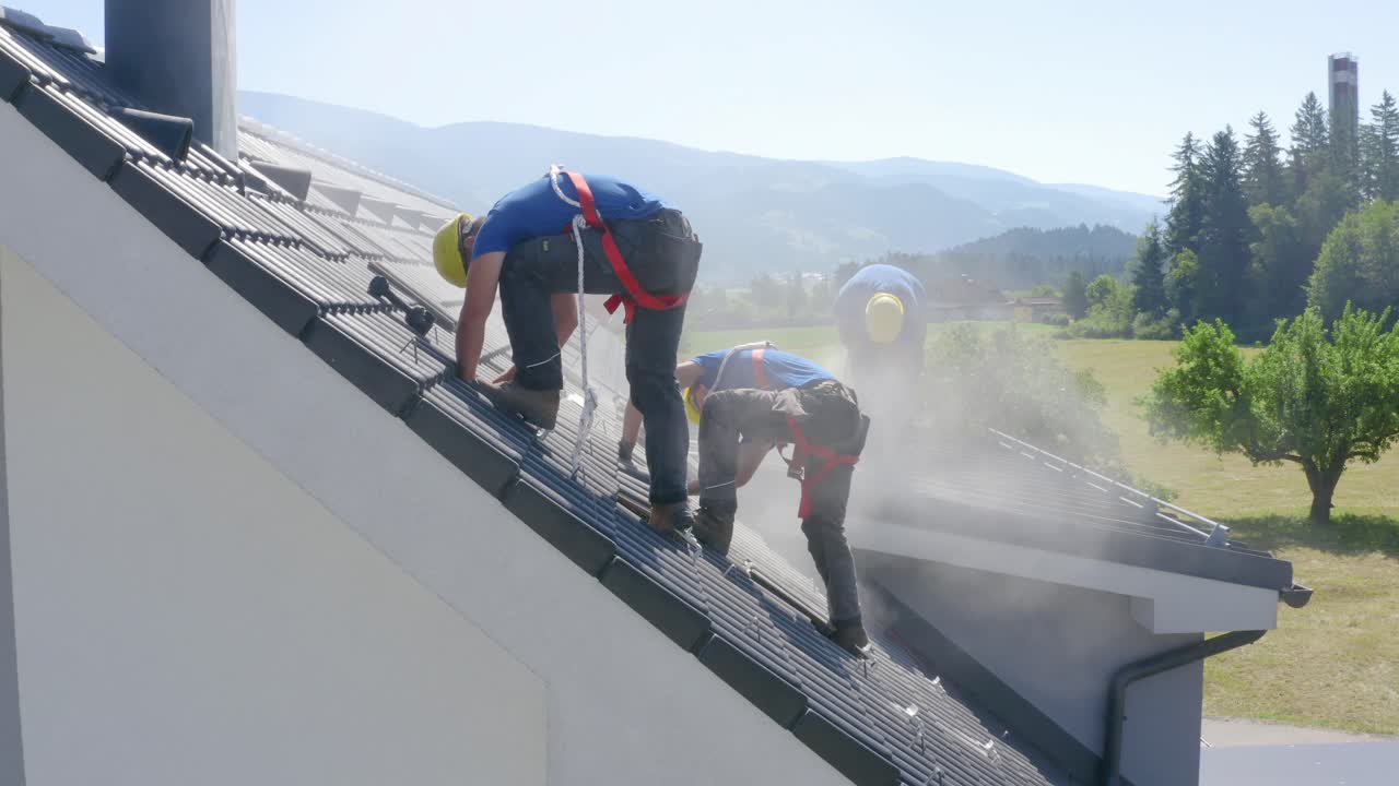 Three young male solar power roof installers with yellow hard hats, blue shirts, and safety harness work and stand on dusty residential rooftop with mountain range in background, aerial