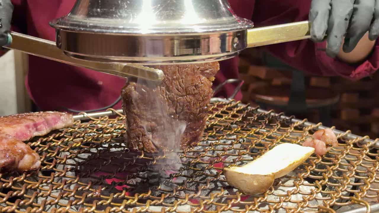 Gloved chef grills wagyu steak with smoke dome on charcoal grill, under warm indoor lighting
