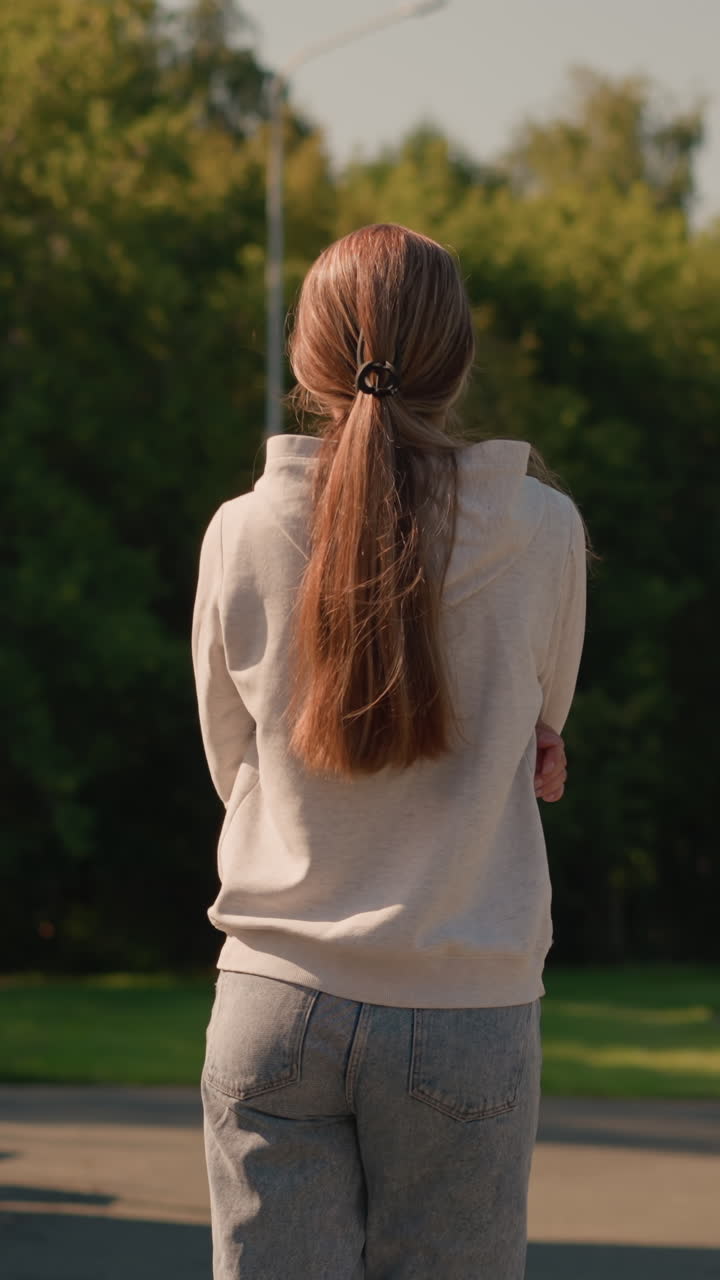 은 날, 전기 기둥과 잔잔한 초록색 나무가 배경에 있는 경기장 좌석 에서 야외를 고 있는 캐주얼 후디를 입은 긴 머리카락을 가진 젊은 여성의 뒷면.