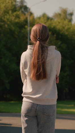 back view of young woman with long hair in casual hoodie, arms folded, walking outdoors by stadium seating on a sunny day, with electric poles and lush green trees in the background