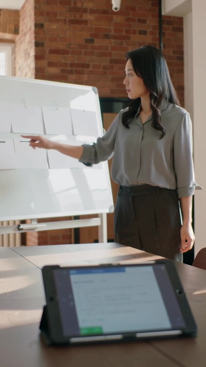 Video verticale: Inizio, donna in top grigio che indica appunti sulla lavagna in ufficio, guidando il team tramite tablet.