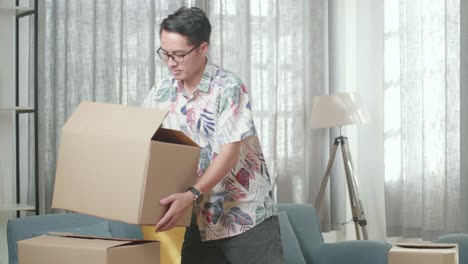 아시안 남자가 휴식을 취하기 위해 소파에 앉기 전에 새로운 집으로 물건을 담은 카드보드 상자를 운반합니다. 그는 새로운 집으로 이사합니다.