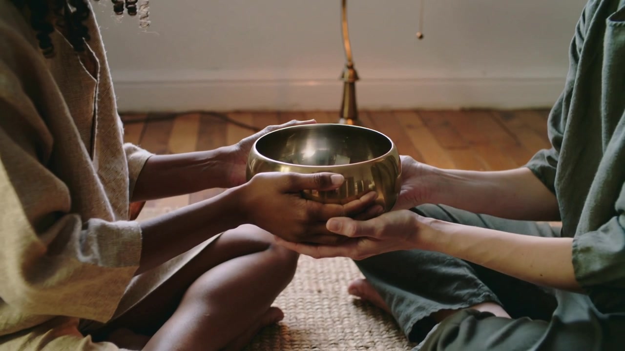 Mani che si protendono, partner che tengono in grembo una ciotola tibetana di ottone in una stanza tranquilla per la meditazione, con una lampada che si accende.