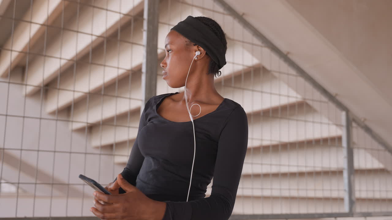 black woman leaning railing checking phone, earphones in, urban stairwell backdrop with concrete geometry, pensive expression, casual black top and purple skirt, scrolling messages while waiting, soft