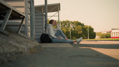 girl in jeans seated on ground with thoughtful expression, resting hand on head, black bag beside her, background features distant trees and building