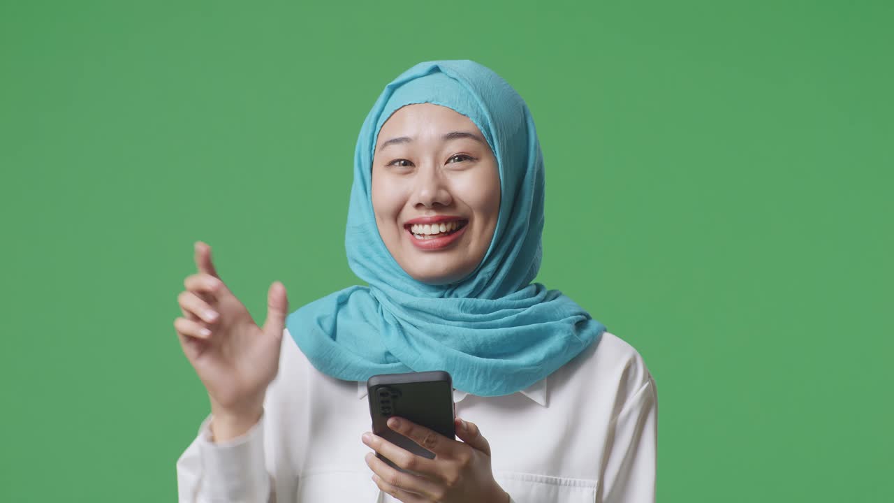 녹색 스크린 배경 스튜디오에 서서 을 가리키는 스마트폰을 사용하면서 미소 짓는 아시아 무슬림 여성의 클로즈업