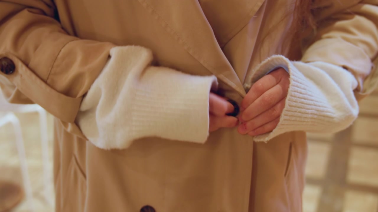 Nahaufnahme einer Frau, die den vorderen Verschluss ihres beigefarbenen Trenchcoats zuknöpft. Die Hände interagieren mit Stoff und Knöpfen, darunter trägt sie weiße gerippte Strickärmel. Weiches Licht erzeugt eine warme.