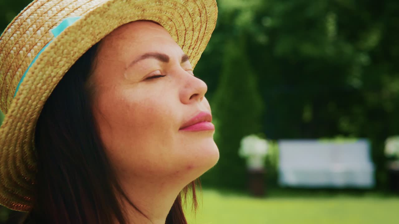 Woman enjoying sunny day outdoors with straw hat and serene expression in natural setting