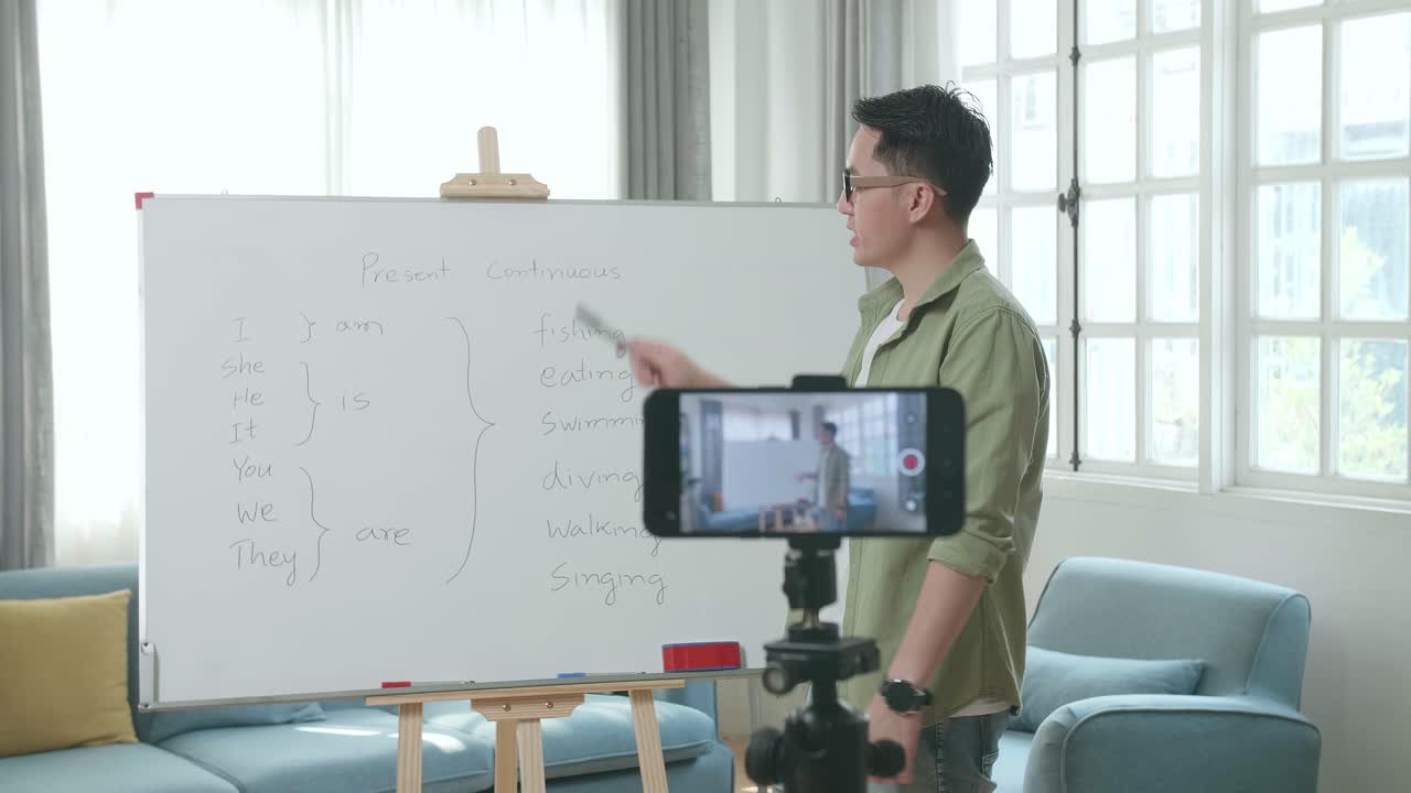 Azjatycki nauczyciel w okularach nagrywający wideo smartfonem, ucząc angielskiego w domu.