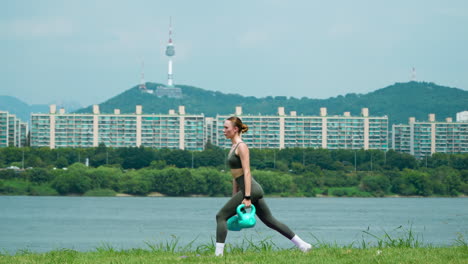 Sportliche-Frau,-Die-Ausfallschritte-Macht-Und-Kesselgewichte-In-Den-Armen-Hält,-Im-Han-River-Park-Mit-Städtischer-Seouler-Landschaft-Im-Hintergrund-–-Outdoor-Workout-Konzept