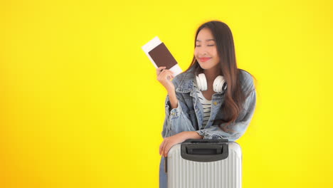 smiling asian woman holding passport and air ticket near suitcase isolated on yellow background