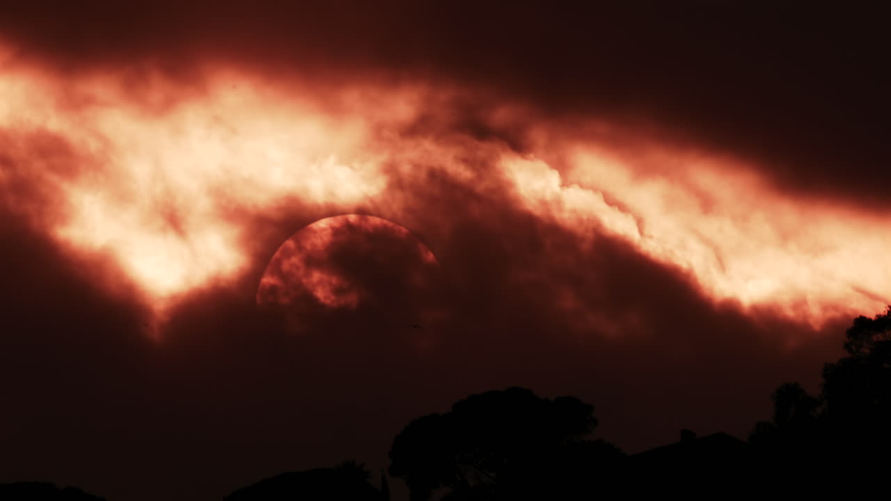 A glowing red sun partially covered by dark clouds at dusk