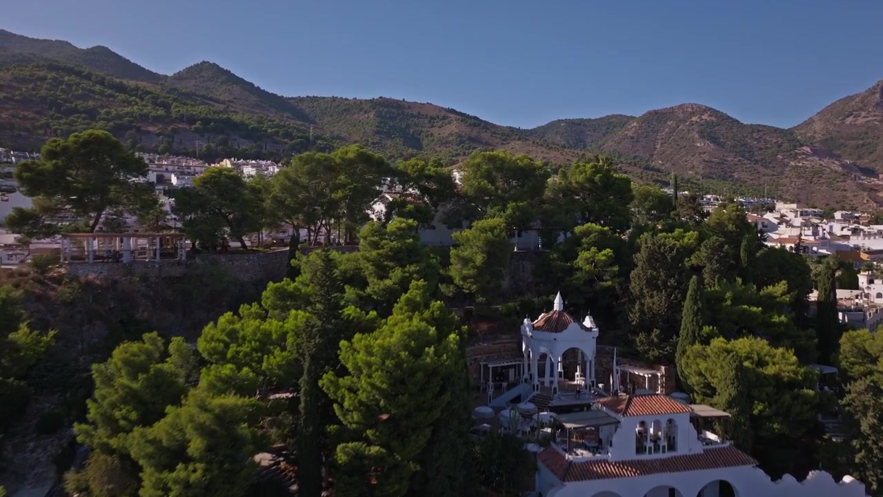 Imagens de drone subindo sobre uma capela arborizada no topo de uma colina em Benalmádena, revelando arcos caiados, pinheiros densos, cristas de montanha e a cidade se espalhando pelo vale abaixo.