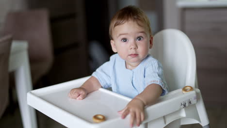 beautiful caucasian baby sitting at the feeding table plays with food. cute toddler moving little bagels by the table and throwing one on the floor.