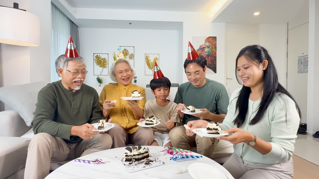 Four generations enjoy birthday cake together in a brightly lit living room, smiling and celebrating