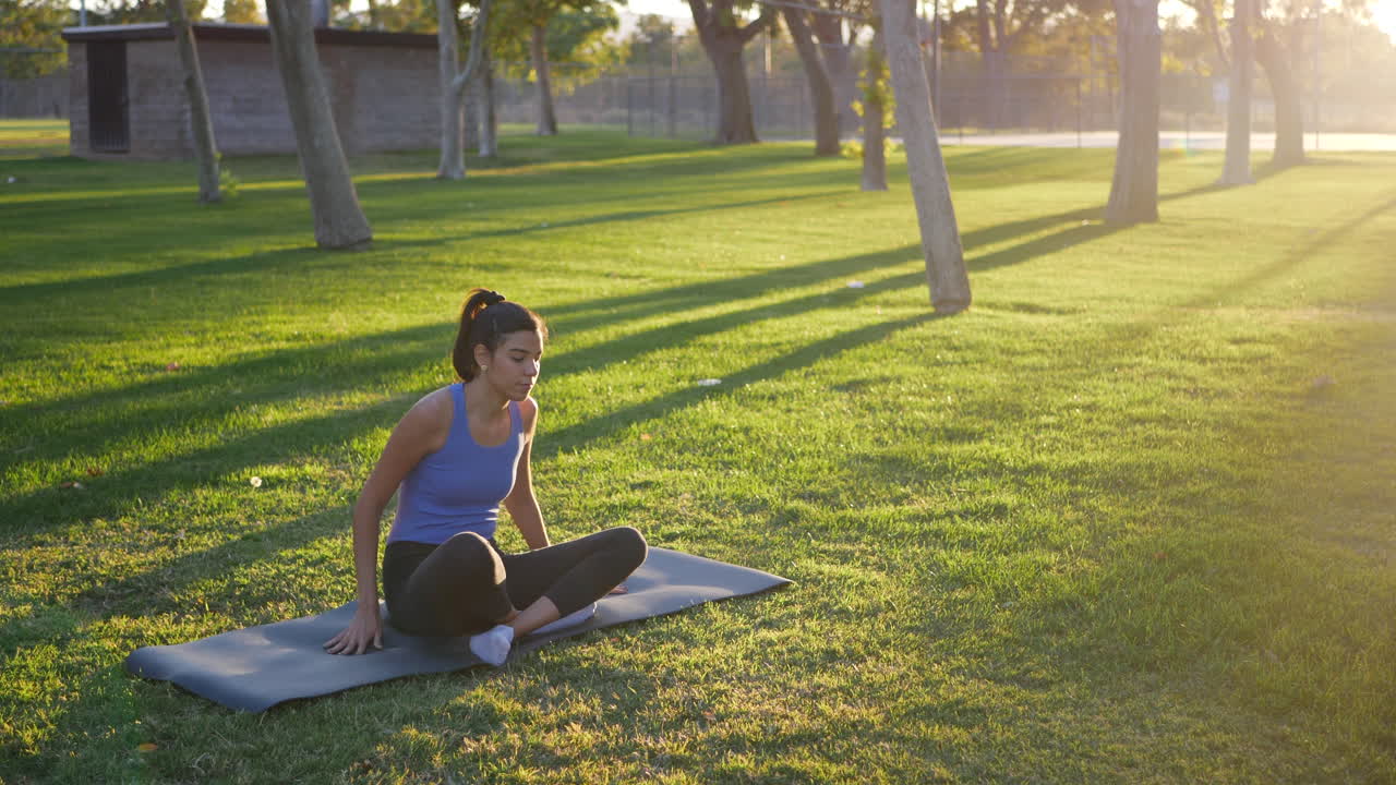 Fit young hispanic woman getting out of lotus position on her yoga mat after a peaceful meditation during sunrise in a grass park