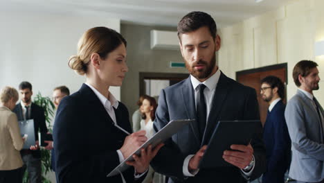 Caucasian-businesswoman-with-a-folder-of-documents-in-hands-talking-and-discussing-something-with-a-businessman-who-holding-a-tablet-during-a-break-in-a-conference-hall