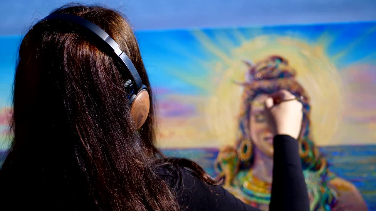 verano, cerca del río, en la playa. al amanecer, una hermosa mujer artista con grandes auriculares marrones sostiene un pincel en la mano, pinta en el lienzo dios shiva. escuchando música, relajarse