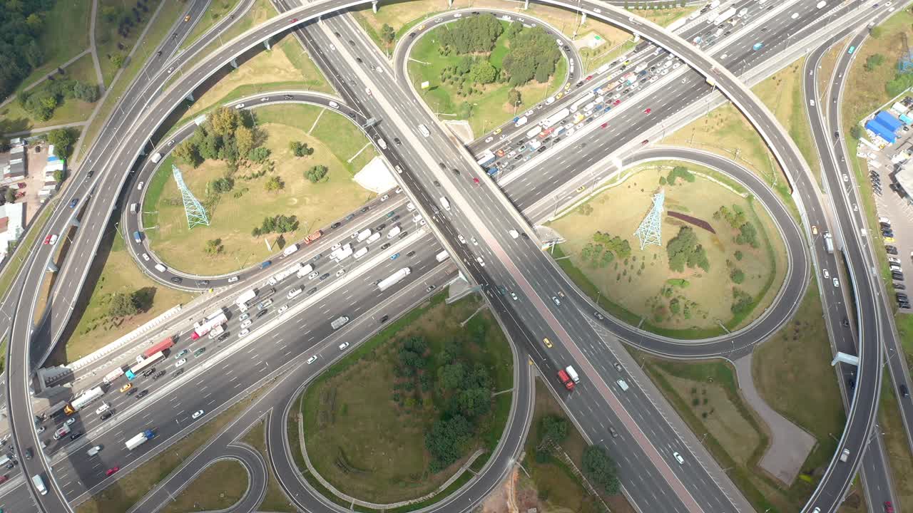 둥글고 구부러진 길과 출구가 있는 큰 교차로, 드론에서 볼 수 있는 최상위 공중 전망, 전형적인 교통 루틴에 많은 자동차, 구름의 그림자와 함께 빛, 잔디와 건물이 있는 초록색 지역, 카메라가 위로, 뒤로 움직이고 있다.