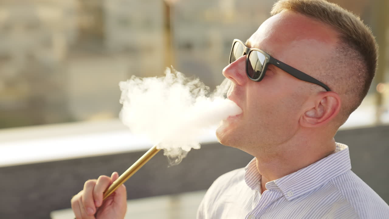 Caucasian young man breathing in smoke from pipe. Male in sunglasses makes a lot of thick white smoke from smoking hookah. Close up.