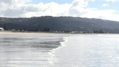 Shallow-wave-flowing-in-diagonally-onto-a-beach-against-a-mountain-background-during-the-daytime