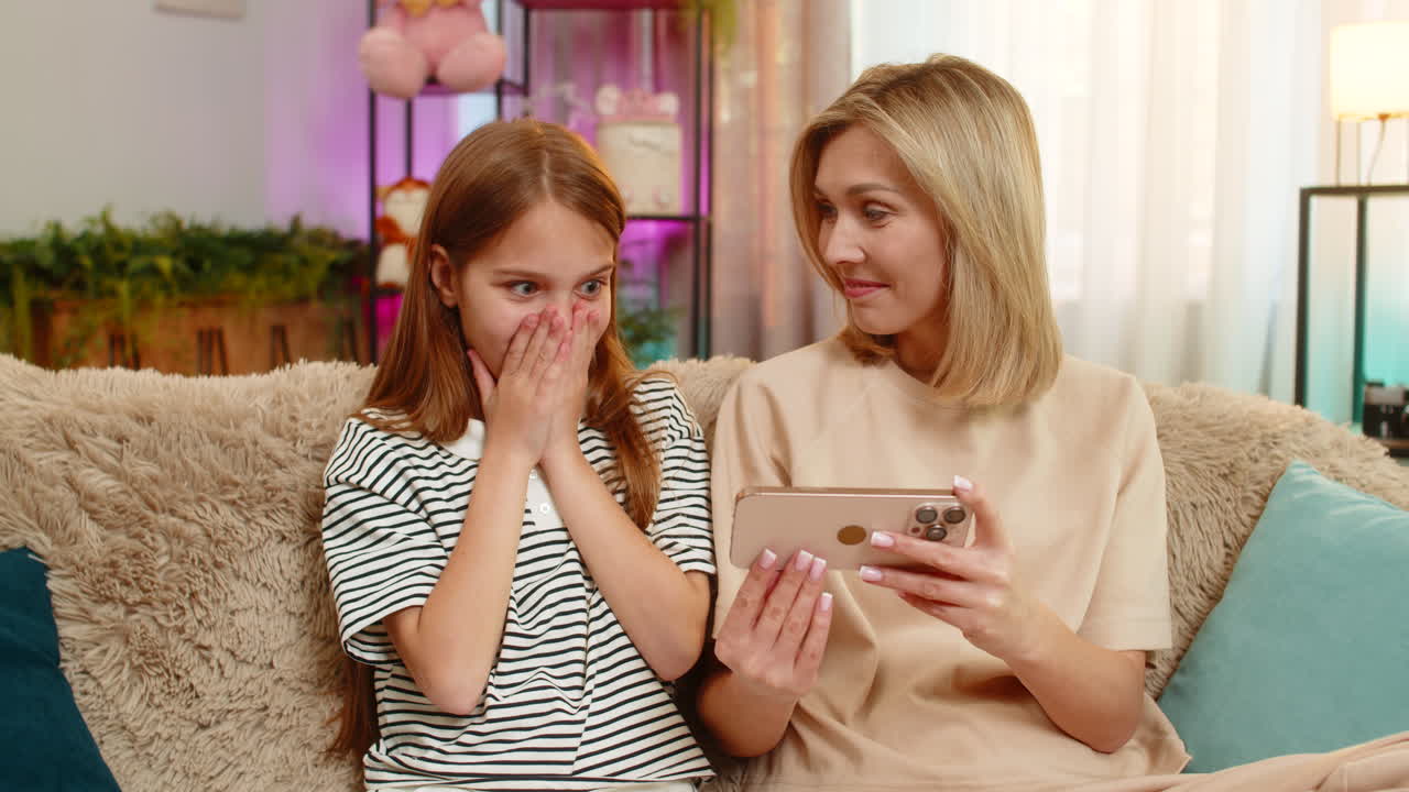 Child girl kid surprised amazed watching wow video on smartphone with mother sitting on home sofa