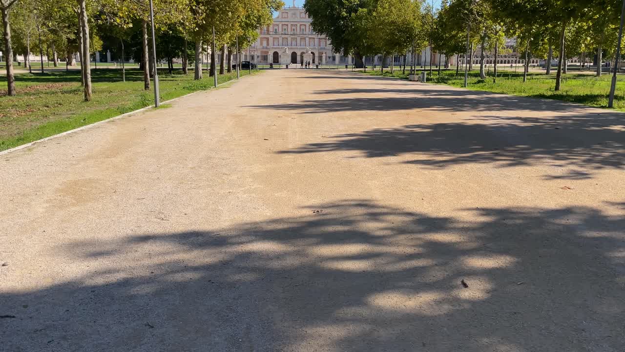 We see in slow motion on the central dirt road of the Royal Palace of Aranjuez where the garden areas are on the sides and with a camera elevation we see the palace in the background and the blue sky.