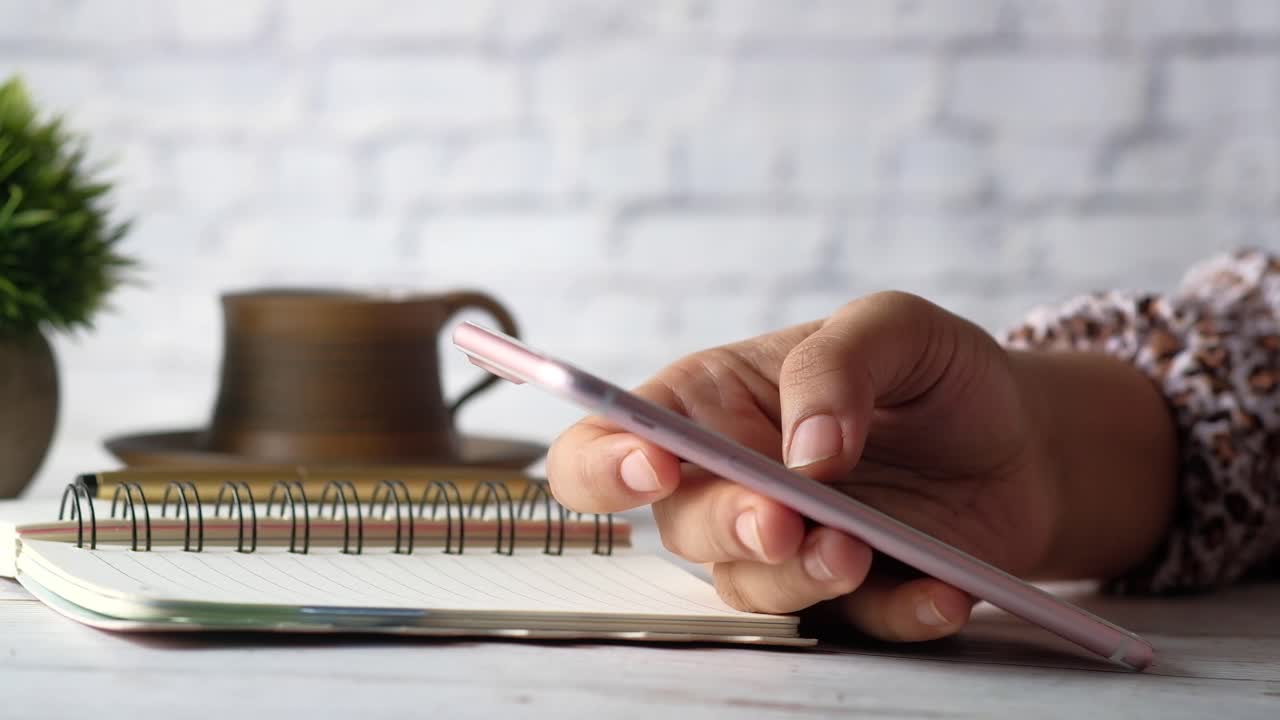 Woman using a pink smartphone at a desk with notebook and coffee cup