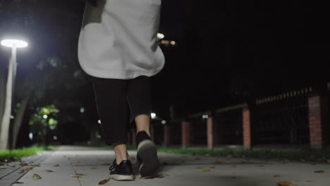 low angle view of woman in white coat walking through park at night with one foot lifted mid-step, dim streetlights casting soft light along quiet path, blurred trees and fence in background