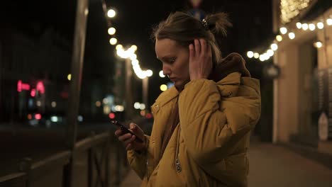 the girl in the jacket is standing on the street with lights in the background. focused at the phone, settle her hair