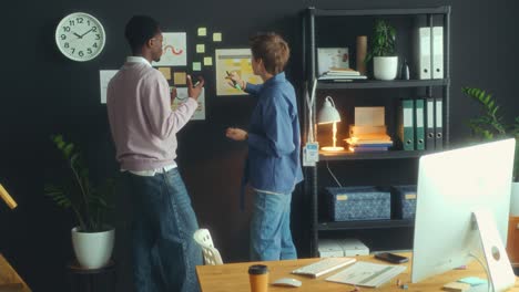two coworkers putting sticky notes on office wall and discussing ideas