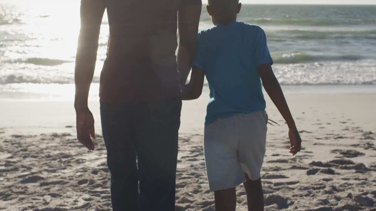 video de un feliz padre y hijo afroamericanos caminando por la playa al atardecer