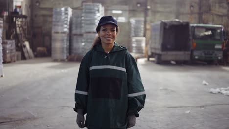 Un-Retrato-De-Una-Mujer-Afroamericana-Con-Un-Uniforme-Verde-Especial-Camina-En-Una-Planta-De-Procesamiento-De-Residuos.-Fondo-De-Fábrica.-Procesamiento-De-Materias-Primas,-Reciclaje.-Control-De-Polución