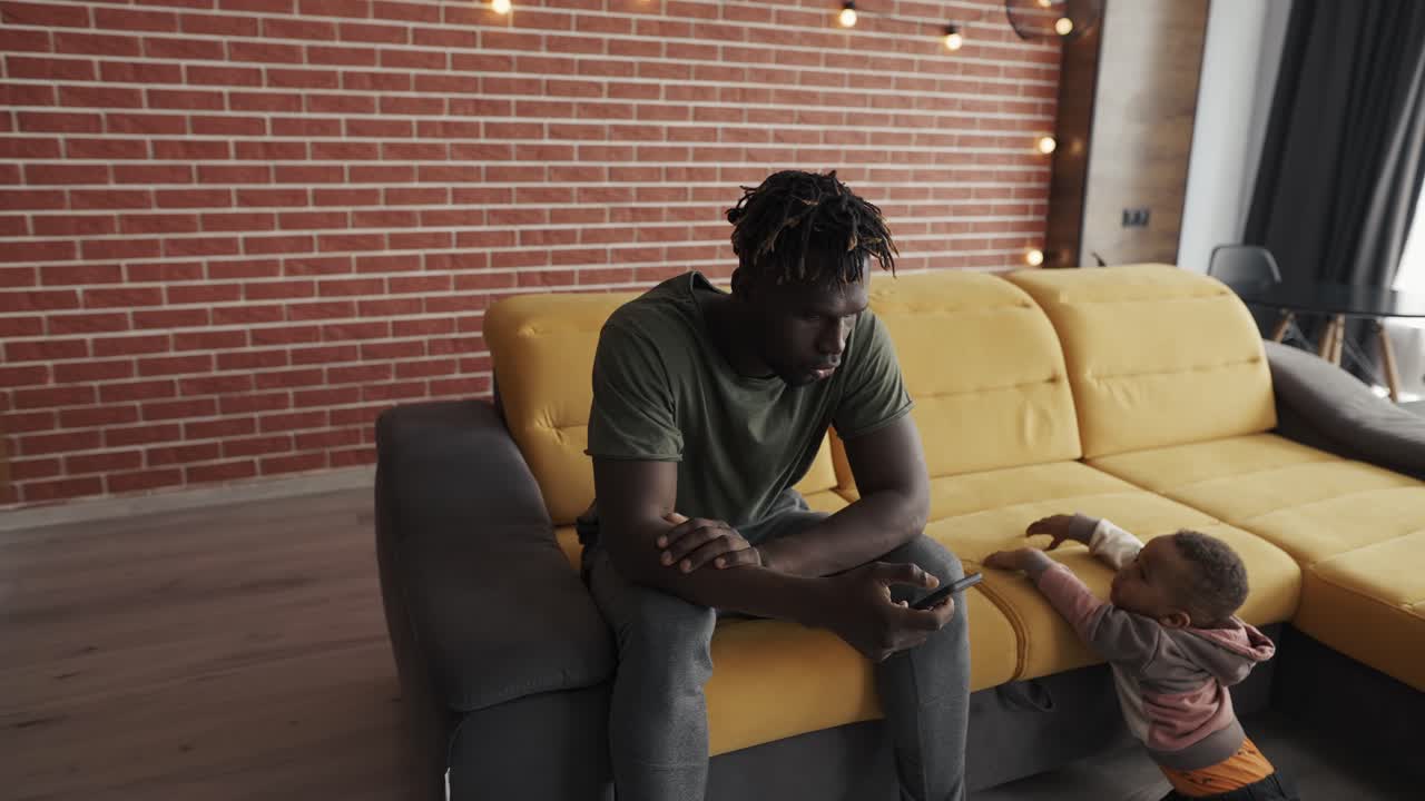 African american young daddy scrolling smartphone while baby son near him bored