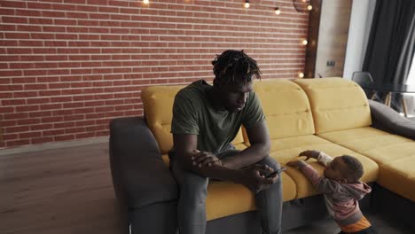 african american young daddy scrolling smartphone while baby son near him bored