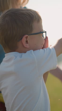 un primer plano de un niño con gafas y una camisa blanca, apuntando al cielo con emoción, mientras su madre sostiene a su hermano menor en el fondo cerca de un lago pacífico