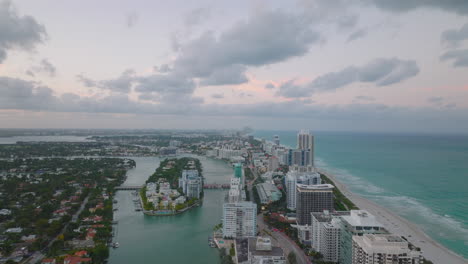 Forwards-fly-above-city-at-dusk.-Modern-multistorey-apartment-buildings-on-stripe-of-ground-between-sea-coast-and-river.-Miami,-USA