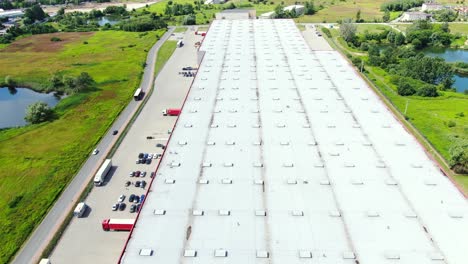 Aerial-Shot-of-Truck-with-Attached-Semi-Trailer-Leaving-Industrial-Warehouse-Storage-Building-Loading-Area-where-Many-Trucks-Are-Loading-Unloading-Merchandise