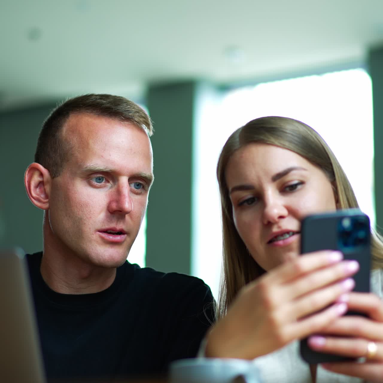Two mid-aged people sitting indoors look at smartphone in lady's hands. Couple talks and discusses something on the phone. Blurred backdrop