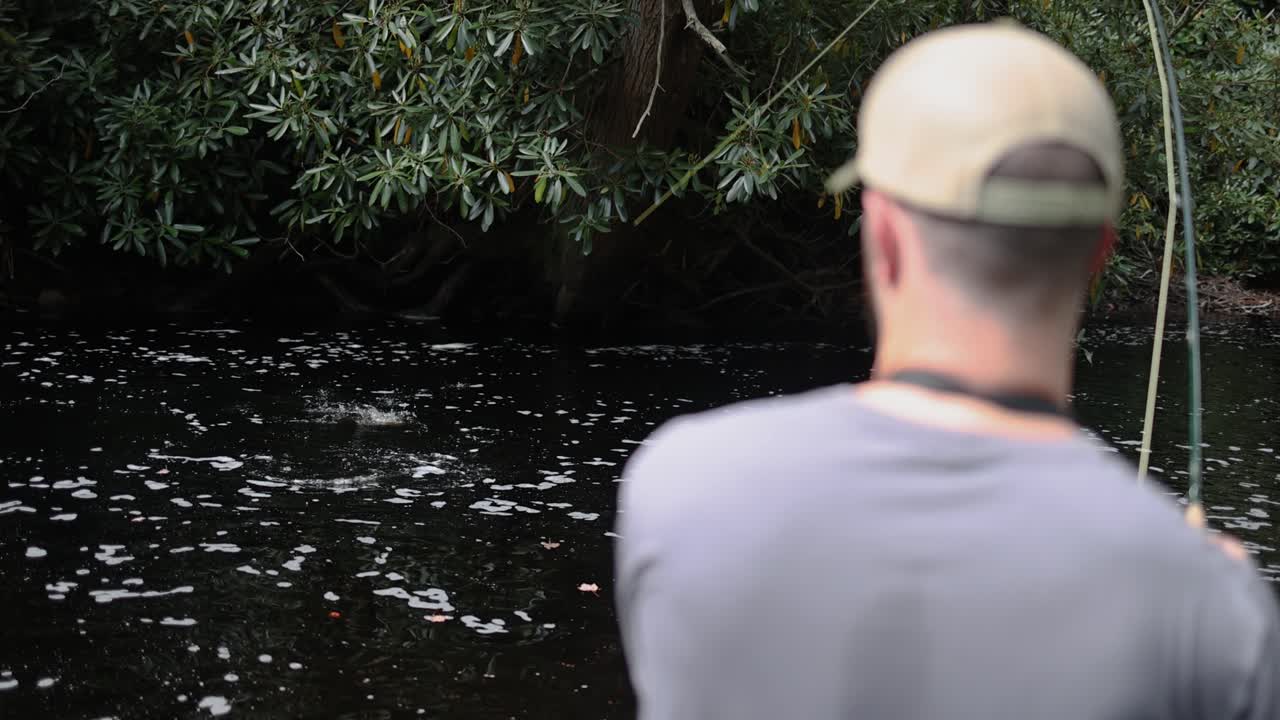 POV over the shoulder slow motion footage of a fly fisherman catching a trout in the stream. The fish is on the line and thrashing in the water. Captured in the Pocono Mountains in Pennsylvania.