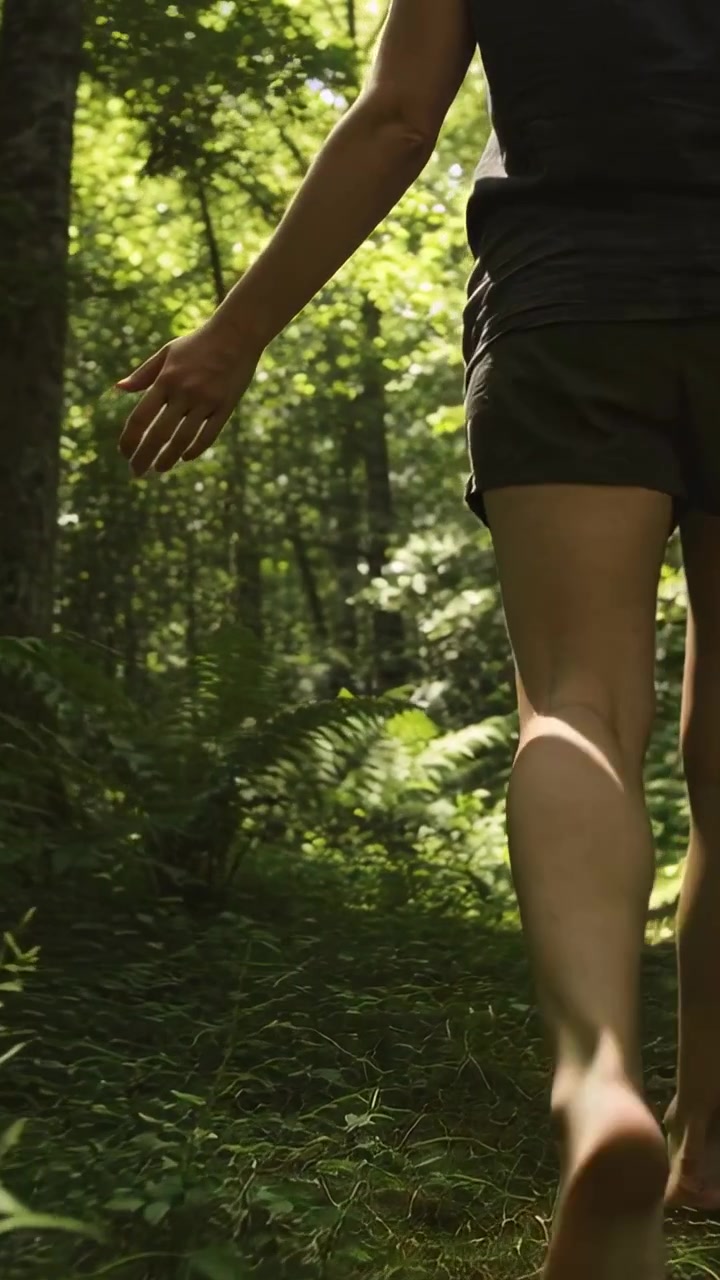 Vertical video: Entering barefoot adult shorts brushing ferns on trail holding sandals, copy space