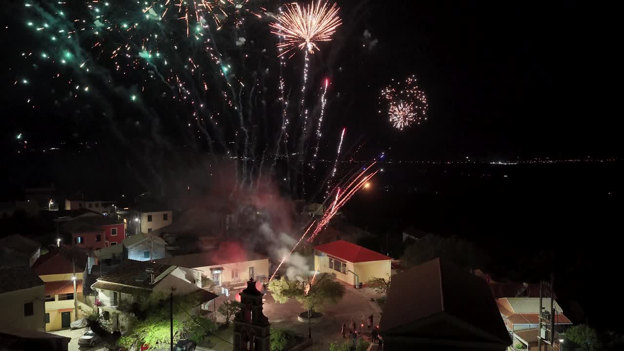 Fantastisk drönarvy av intensiva, färgglada fyrverkerier som exploderar direkt över en kyrka och hus i en traditionell by på Korfu, Grekland, på natten.