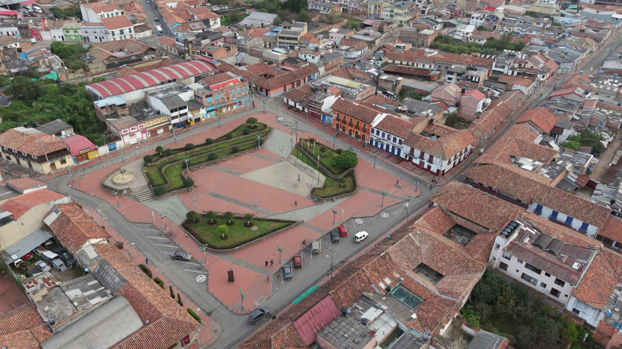 Drone footage captures Plaza de la Independencia, Zipaquirá, showcasing lush gardens and colonial architecture in vibrant contrast under a soft overcast sky. A captivating urban landscape