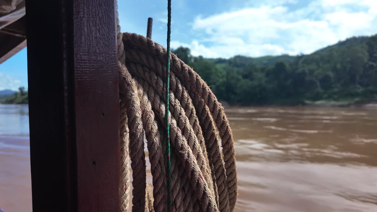 Corda arrotolata su una barca di legno accanto al fiume Mekong in Laos, con lussureggianti colline verdi. Rallentatore