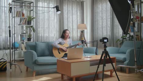 mujer asiática con una computadora portátil agitando la mano y hablando a la cámara mientras transmite en vivo tocando una guitarra en casa