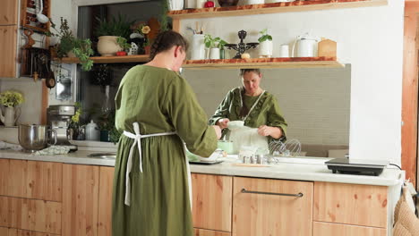 woman in olive green dress preparing breakfast in cozy rustic kitchen with full mirror reflection of her actions on clean counter, surrounded by modern appliances, herbs, and utensils